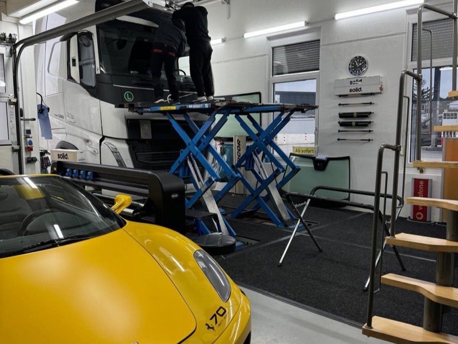 LKW-Frontscheibe auf dem Scherenhubtisch, daneben ein Sportwagen